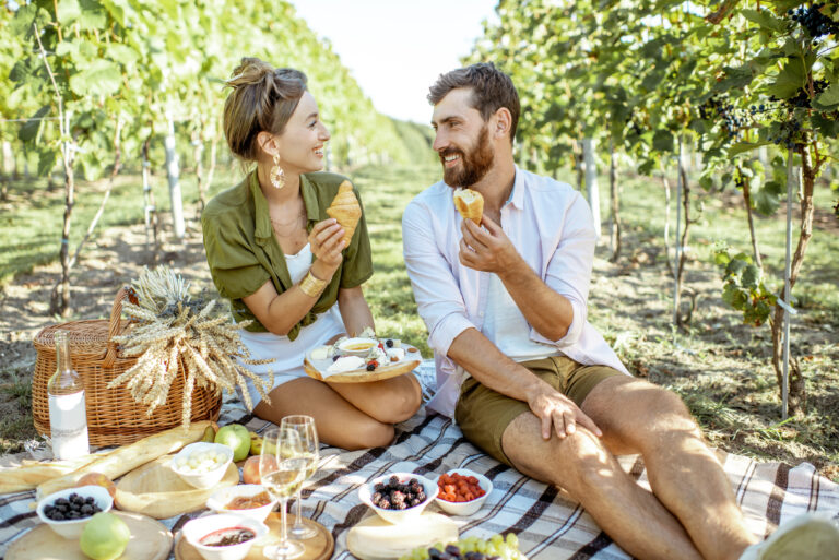 Beautiful couple having romantic breakfast with lots of tasty food and wine, sitting together on the picnic blanket at the vineyard on a sunny morning